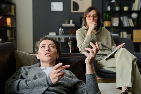 Woman lying on couch talking while therapist listens and takes notes in background Communicating thoughts and emotions during therapy session in calm, supportive environmentの写真素材