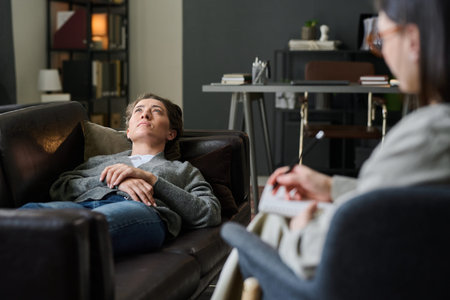 Individual lying on couch during therapy session in cozy modern office decorated with bookshelves and plants reflecting on thoughts and emotionsの写真素材