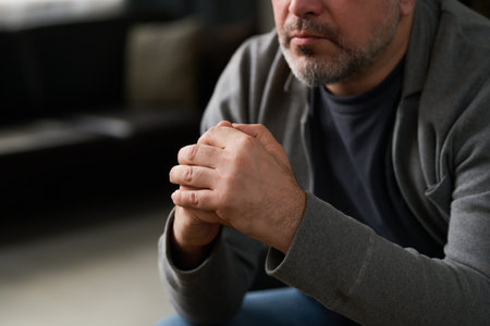 Caucasian man with short beard and folded hands contemplating in intimate indoor setting with thoughtful expression and detailed background elementsの写真素材