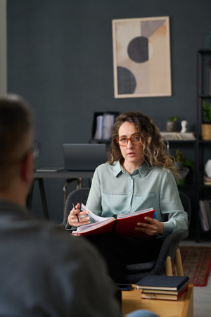 Professional woman holding open notebook engaged in conversation with colleague in modern office setting, with wall art and shelves in backgroundの写真素材
