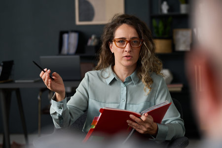 Therapist consulting senior patient in modern office, holding pen and notebook recording session. Background shows organized bookshelf and potted plantの写真素材