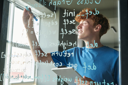 Side view of red haired man writing computer programming code on glass wall during IT class in schoolの写真素材