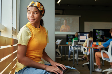 African American young woman using laptop while sitting by window in class at school or college and...
