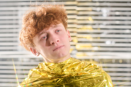 Young man standing with gold reflective material around shoulders, facing away from camera in brightly lit environment with striped background creating unique light patternsの写真素材