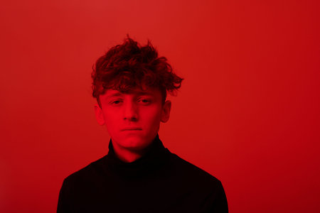 Portrait of young man with curly hair looking directly at camera, illuminated by red light, creating...