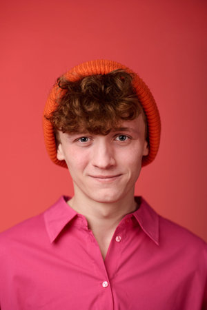 Portrait of young Caucasian man smiling and wearing a bright orange beanie and pink shirt against a...