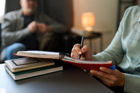 Hand writing notes in red notebook during counseling session with client in blurred background...
