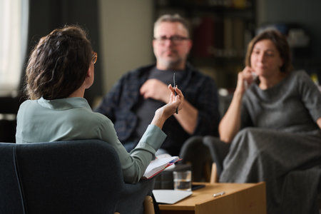 Couple having therapy session with female therapist gesturing while holding pen, room with bookshelf and therapy materials, focus on conversation, atmosphere calm and professionalの写真素材