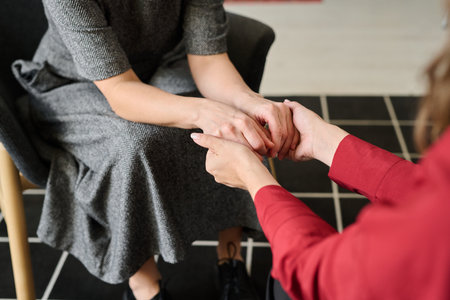 Comforting scene showing supportive gesture by two people sitting indoors with one person gently holding hands of the other, offering reassurance and warmthの写真素材