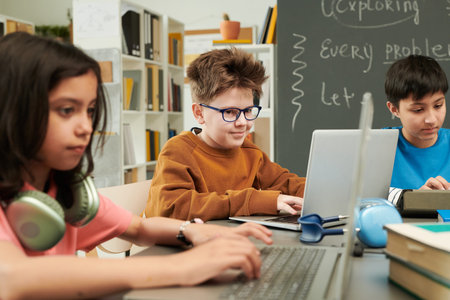 Portrait of young boy with funny hair and glasses using laptop with group of children at table in...