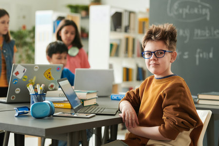 Portrait of funny young boy wearing glasses looking at camera in school library during computer...