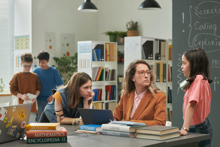 Portrait of female teacher talking to girls during computer class in school library copy space
