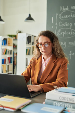 Vertical portrait of female teacher wearing glasses and using computer while working at desk in...