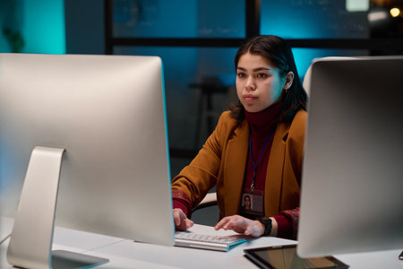 Portrait of successful young woman as female cybersecurity expert using computer in office and looking at screen with blue lightの写真素材