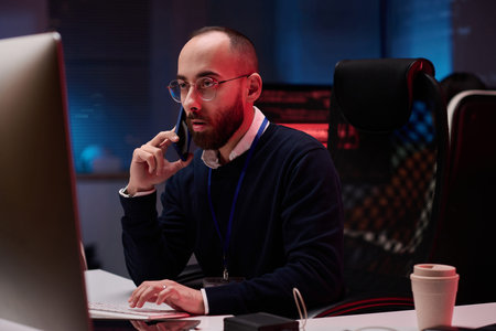 Portrait of adult man using computer and speaking on phone while working on emergency in...