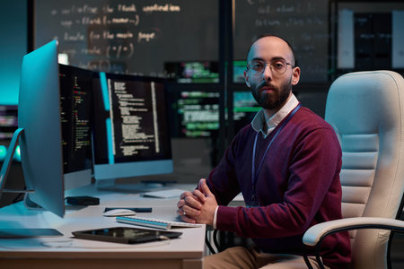 Portrait of bearded network security engineer looking at camera sitting at workplace with code...