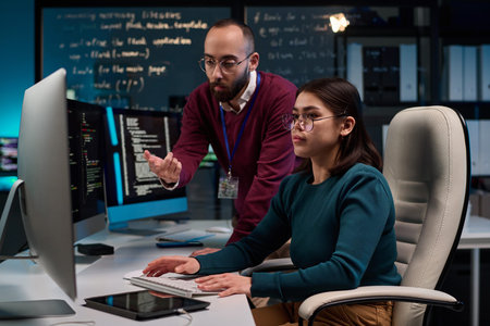Portrait of young woman using computer in software development office with senior mentor giving...