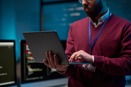 Close up of bearded adult man holding laptop standing in IT development office with blue lights copy...