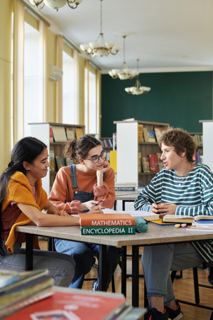 Group of young students sitting around a table in a library, engaging in collaborative study session with books and notebooks. Bright light fills the room, enhancing the studious atmosphereの写真素材