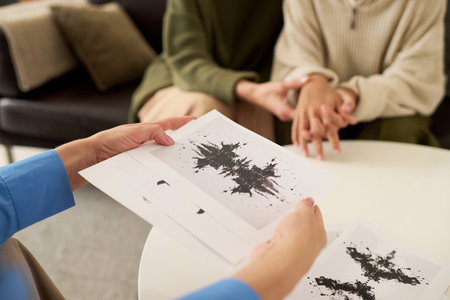 Female therapist holding inkblot test in hands with teenage girl and mother sitting together holding hands in background, mental health assessment session, copy space.の写真素材