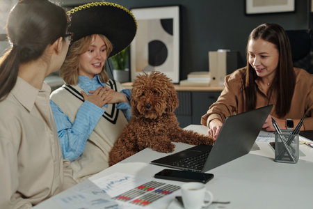 Candid shot of young women enjoying work together in office and chatting cheerfully with girl wearing sombrero hatの写真素材