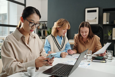 Portrait of young Asian woman using smartphone and checking social media during business meeting with all female team in officeの写真素材