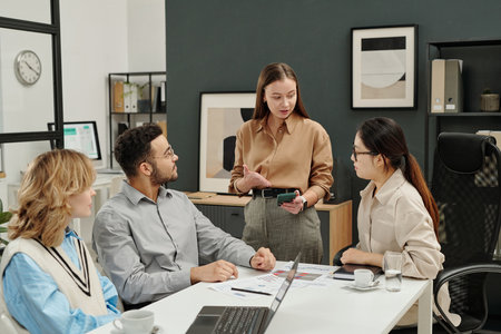 Portrait of young businesswoman as female boss pointing at smartphone screen while discussing marketing in business meeting with team in officeの写真素材