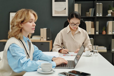 Candid shot of two young businesswomen coworking in open office setting busy with individual tasks and using laptopの写真素材