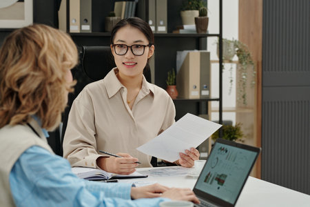 Portrait of young Asian businesswoman smiling while holding document and talking with female colleague in business meeting at modern officeの写真素材