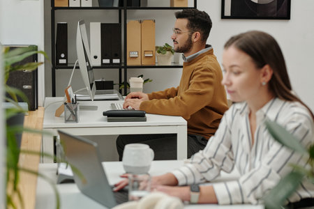 Side view portrait of young man using computer while sitting at desk in modern office decorated with live plants female colleague typing on laptop keyboardの写真素材