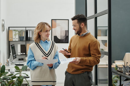 Waist up portrait of two young people standing in office and holding documents while discussing work issuesの写真素材