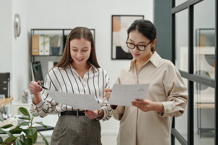 Waist up portrait of two smiling young women holding documents and discussing data while standing in modern office settingの写真素材