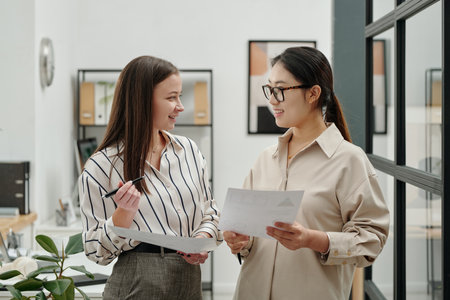Side view portrait of two smiling young businesswomen looking at each other while discussing documents standing in modern office settingの写真素材
