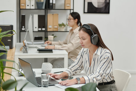Side view of smiling young woman wearing headphones while typing on laptop in modern office with colleagues working in backgroundの写真素材