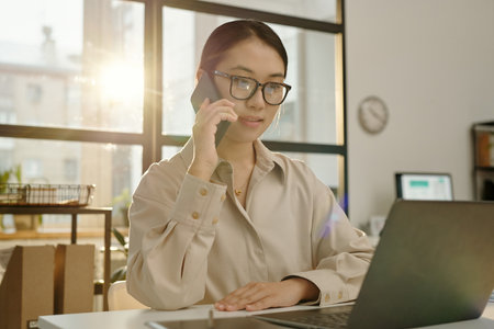Portrait of young Asian woman speaking by phone and using computer while sitting at desk lit by sunlight glow copy spaceの写真素材