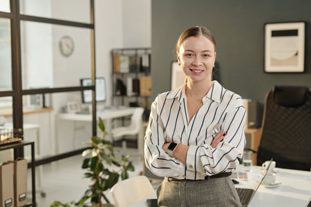 Waist up portrait of confident young businesswoman standing with arms crossed in modern office and...