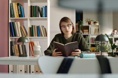 Portrait of female student reading book and wearing glasses while sitting at desk in college library...