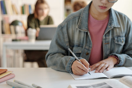 Close up of young woman as female student writing in notebook while studying at desk in school or...