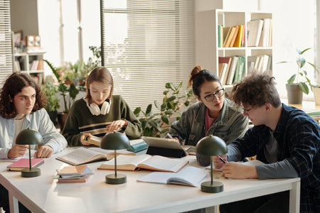 Group of college students collaborating and studying together at desk in classroom with books and stationary scattered on table scene lit by sunlightの写真素材