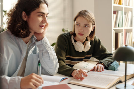 Portrait of young girl looking at friend while studying together at desk and reading open book on tableの写真素材