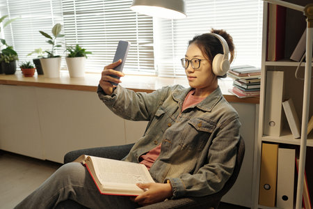 Side view portrait of young Asian woman taking selfie photo in school library and wearing headphones...