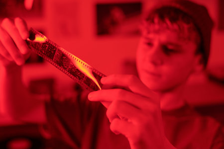 Close up of young man holding film roll to light while working on analog photography in darkroom lit by red light copy spaceの写真素材