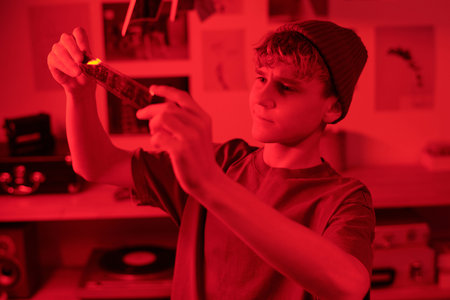 High angle view at focused teenage boy looking at developed film selecting photos for printing against red neon light in darkroom copy spaceの写真素材