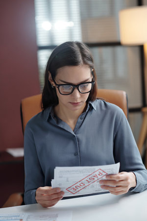 Vertical portrait of young woman holding bills with past due stamp while sorting financial documents in office or filing taxesの写真素材