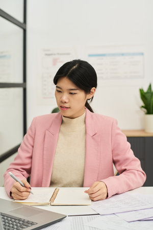 Asian woman sitting at desk, taking notes and studying foreign languages in classroom environment with charts and plant in background, engaging with open notebookの写真素材