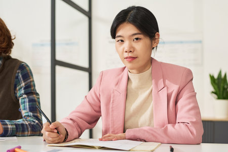 Asian woman attentively participating in language learning class while holding a pen, wearing pink blazer, and seated at desk with study materialsの写真素材