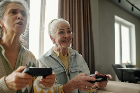 Two senior women sitting in living room engrossed in playing video game together, holding...