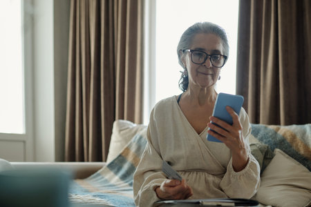Elderly woman sitting on couch while engaging with smartphone and remote control, wearing eyeglasses...