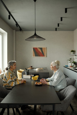 Elderly women conversing over fruit at modern dining table with abstract art on the wall sharing an intimate and meaningful momentの写真素材