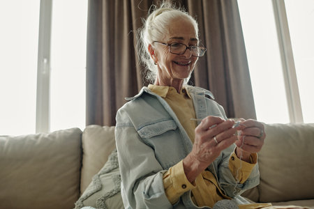 Senior woman sitting on sofa while holding a piece of yarn and knitting. She is wearing glasses and a light jacket, bathed in soft indoor light with a serene expressionの写真素材